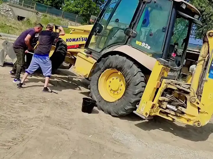 Buldoexcavator galben ridicat pe cric, cu doi mecanici la roata din față
